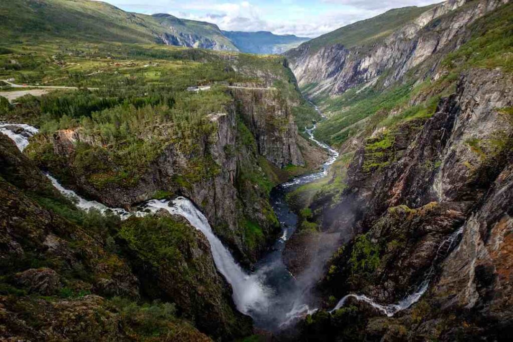 A famous waterfall in Norway beautifully cascading with whitewater down the mountain sides into a ravine, amidst green summer mountains stretching into infinity.