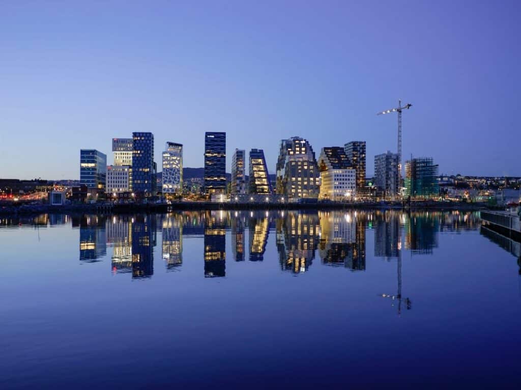 Beautifully lit Barcode Skyline of Oslo Norway at night. The sea is blanc and calm in the front, mirroring the entire skyline, in front of a deep blue sky. 