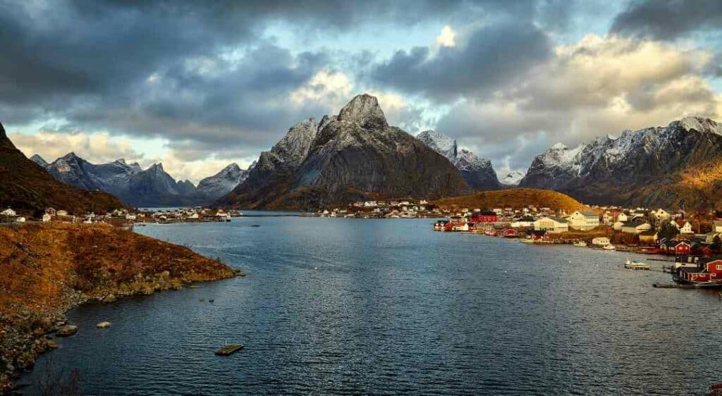 A small village in Lofoten Norway, with the rugged fjord in the front, with patches of sun over the nature, against a spectacular backdrop of the Lofoten mountains with snowy peaks in the background. 