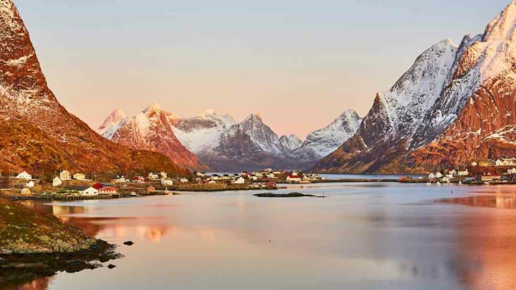Incredible scenery with the blank fjord in the foreground, a small village with red and white wooden houses in front of majestic mountains with snow covered peaks in the low glowing winter sun