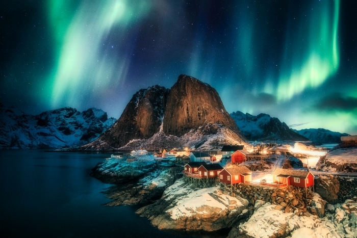 Northern lights in waves of green on the starry night sky in Lofoten Norway, with a small community of red wooden houses in the front on the snowy shores. 