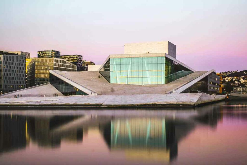 The Oslo Opera hours seen from the fjord, with a pale sky in the background, and the winter sun setting leaving a greenish light in the large glass facade, and a pink shine on the blank water