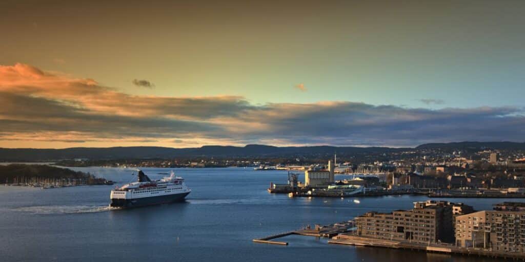 A cruise ship sailing towards port in Oslo, in the inner harbor with modern buildings along the shore, at sunset with a glowing light in the light blue sky