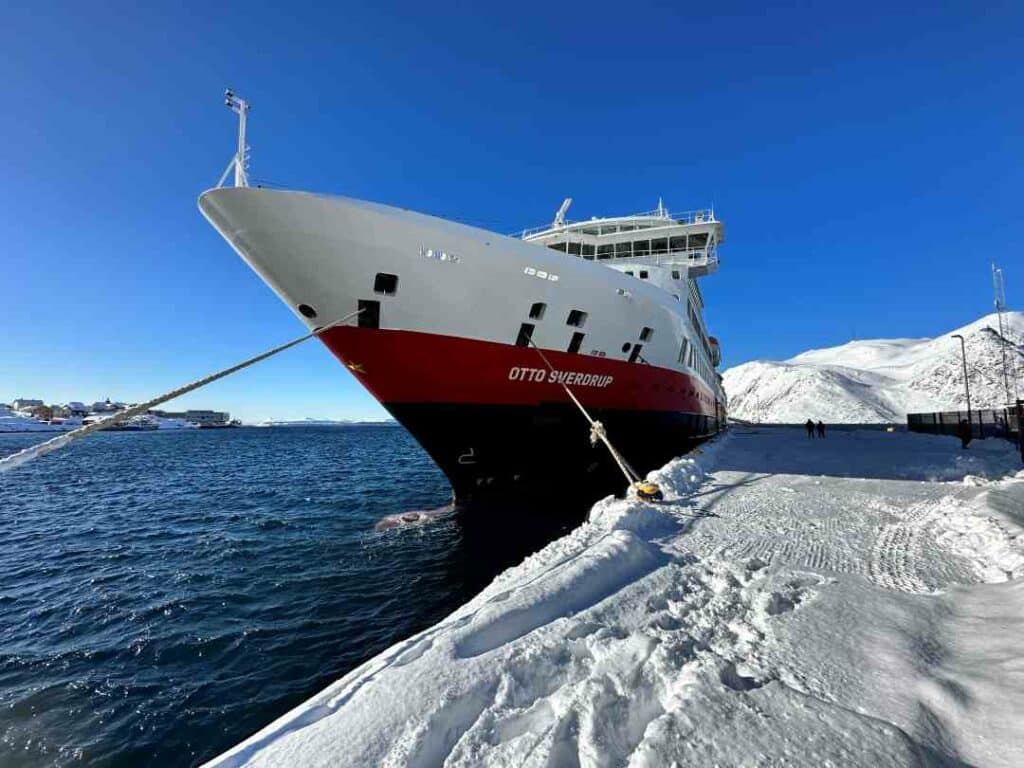 Otto Sverdrup white and red ship docked in Honningsvåg close to the North Cape in the north of Norway on a bright winter day. The snow is crispy white, the sea deep dark blue, and the sky incredibly bright blue, with white mountains in the distance. 