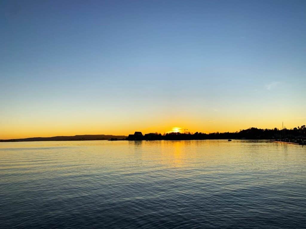 The Bygdøy Peninsula across the dark fjord during sunset with a glowing sun in the distance reflecting in the fjord