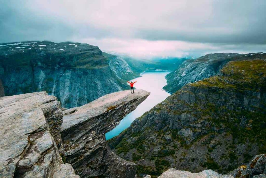 Spectacular views from Trolltunga in Norway. A small person in a red coat standing on the Trolltunga mountain cliff high above the silvery fjord below, with steep mountains  on either side and snow capped mountains disappearing into the distance. 
