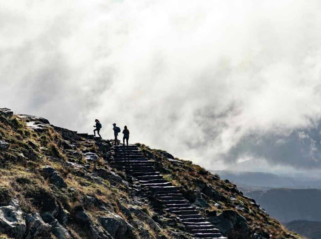 Three hikers walking up the Ulriken mountain in Bergen, Norway. The cloud cover is flowy and thin, and the sun is sifting through. In the background you see more hilly mountains. 