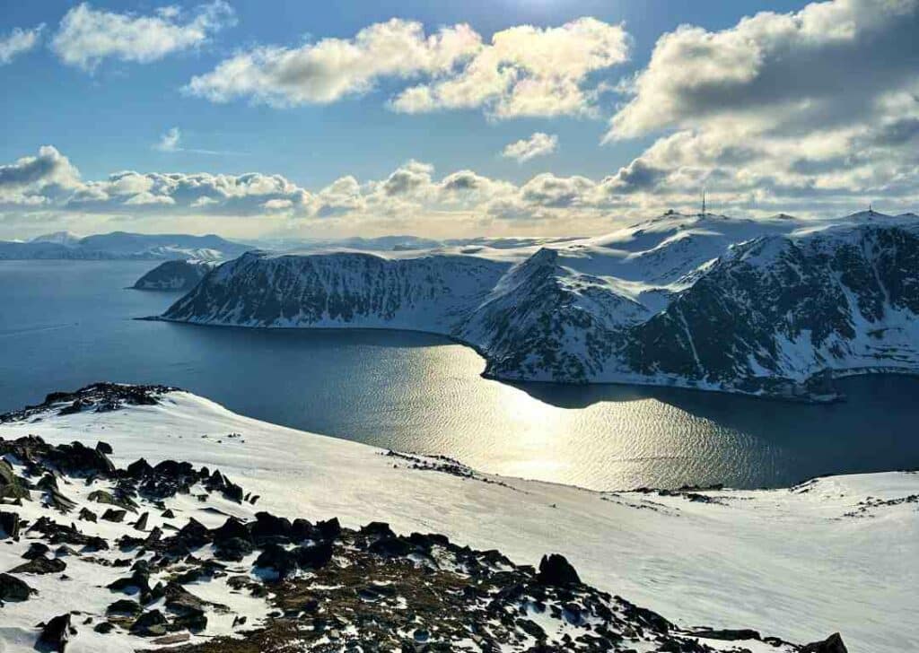 View from the summit above the Sherpa Stairs in Honningsvåg, spectacular views of the fjords and the vast snow covered mountains that have steep sides straight into the fjord on a bright sunny day with incredible reflections of the sun in the deep blue water. 