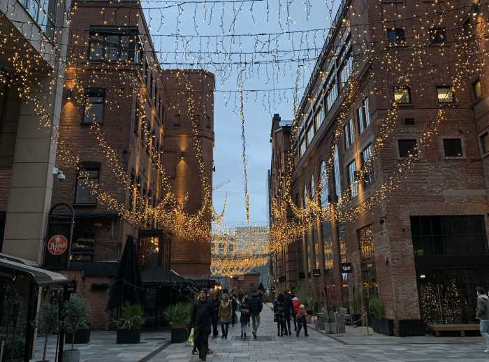 Aker Brygge decorated for Christmas with light girlanders hanging between the maroon brick buildings, and you can see the pale blue winter sky above the buildings