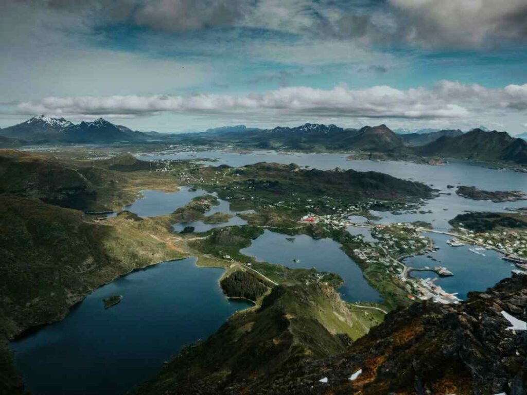 Stunning views of Ballstad, Lofoten, Norway from a mountain top, with infinite views of the islands and the sea under a partly cloudy sky and snow patched mountains in the distance