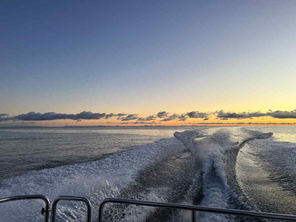 Boat trip at sunset in Arctic Norway, where the sun is right below the horizon, creating a golden stripe of light between the dark silvery sea and the soft pale blue sky. 