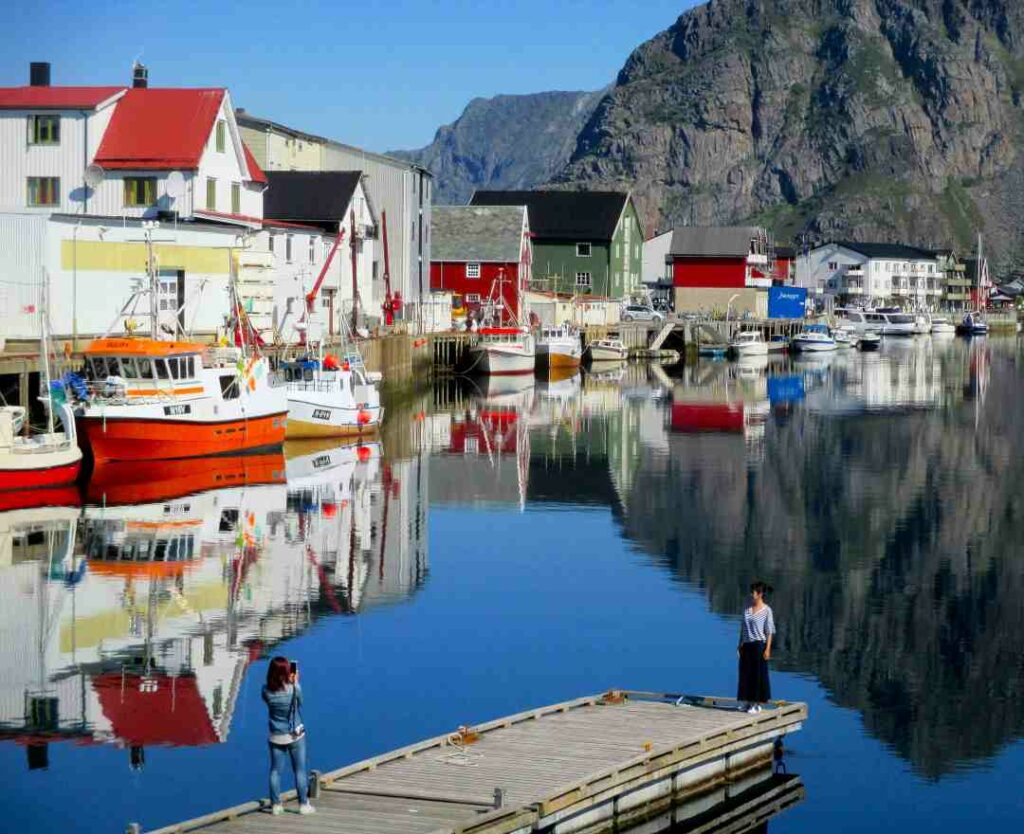 Henningsvær fishing village on a bright sunny summer day, with colorful houses and fishing boats in the foreground, and the majestic mountains in the background under the blue skies. 