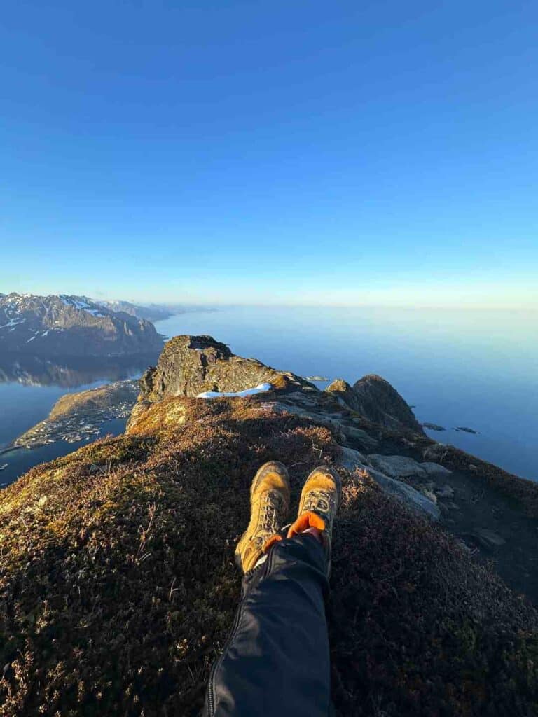 A hikers booted feet on top of a mountain ridge sitting on green moss, with spectacular views to the  deep blue fjord and sea below, majestic mountains in the distance, under a blue sky in the Lofoten Islands Norway in the spring