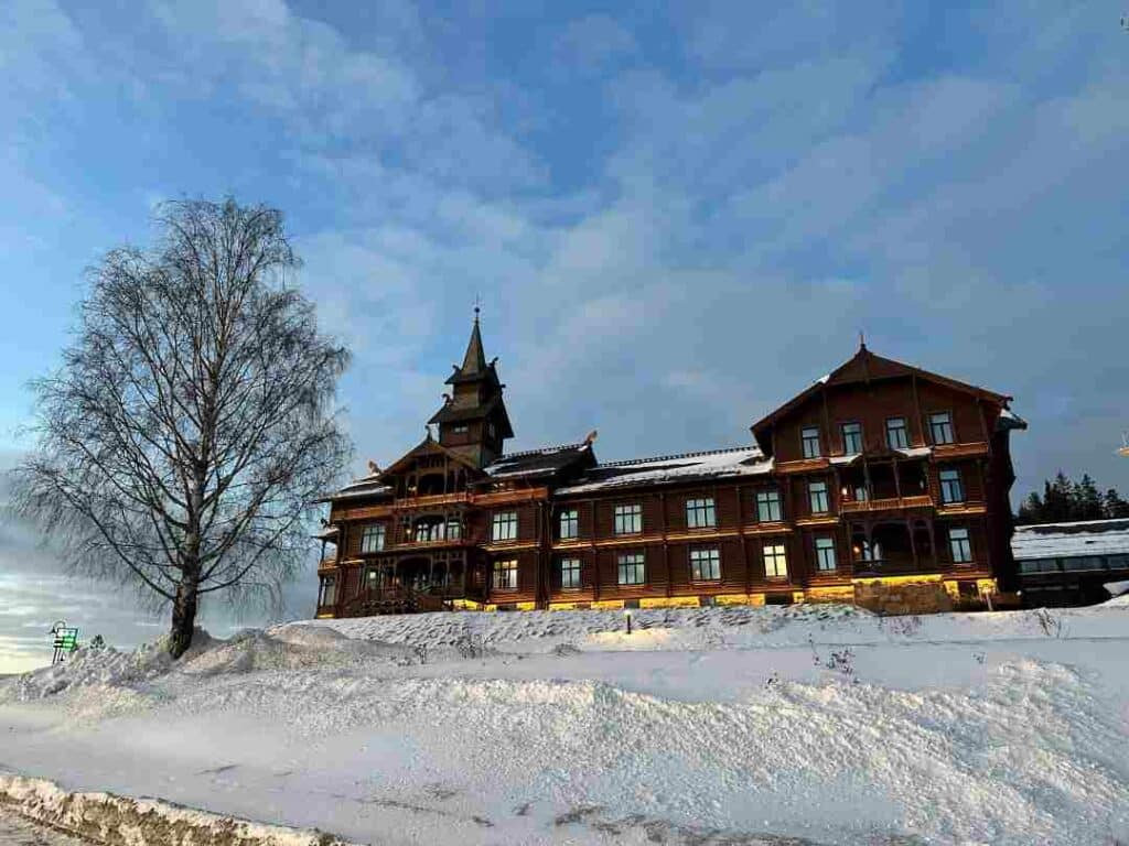 The venerable Holmenkollen Hotel above Oslo, with its impressive wooden dark facade, with decorations, traditional windows, multi-layered roofs and speer on a sunny and cold winter day with crispy white snow, blue skies and a winter tree without leaves. 
