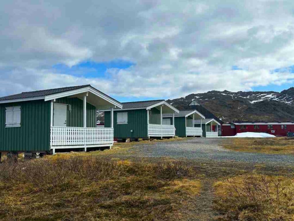 Small green camping cottages with white porches on the tundra with yellow grass, and mountains with snow patches in the background