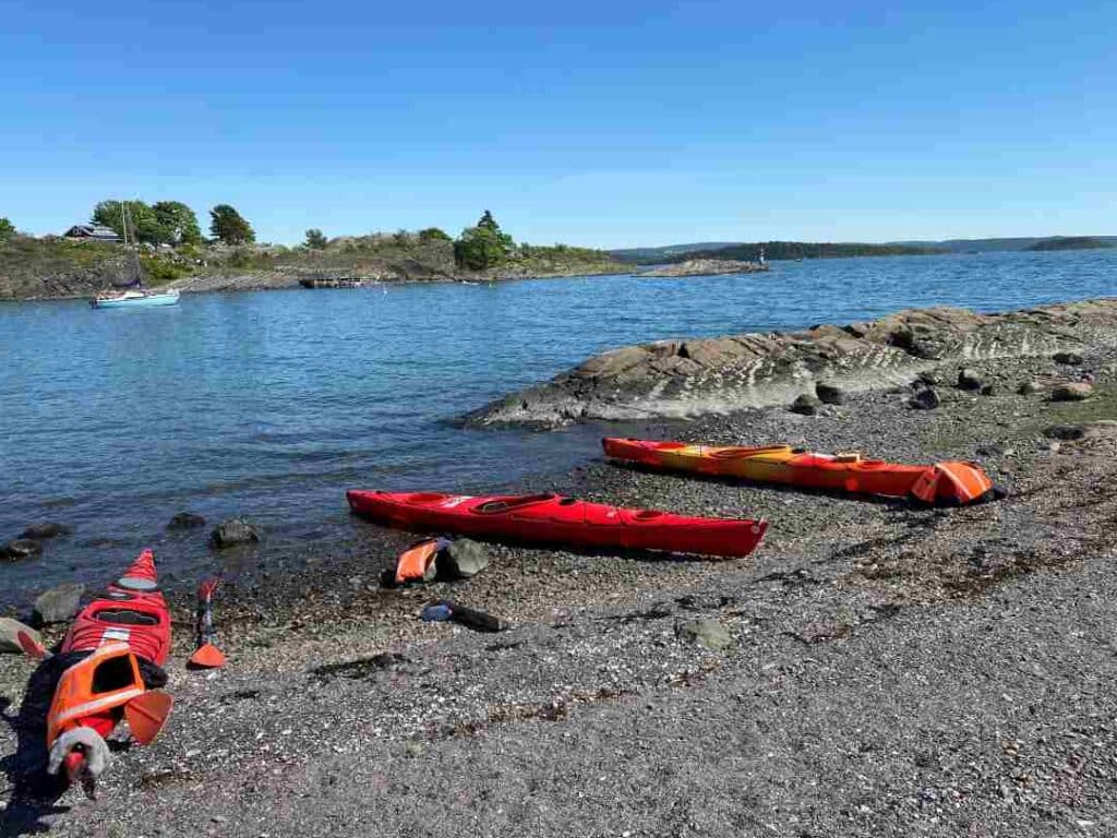 Three red kayaks lying on a pebbled beach with rocky shores to a deep blue fjord on a summer day bathed in sunlight. 