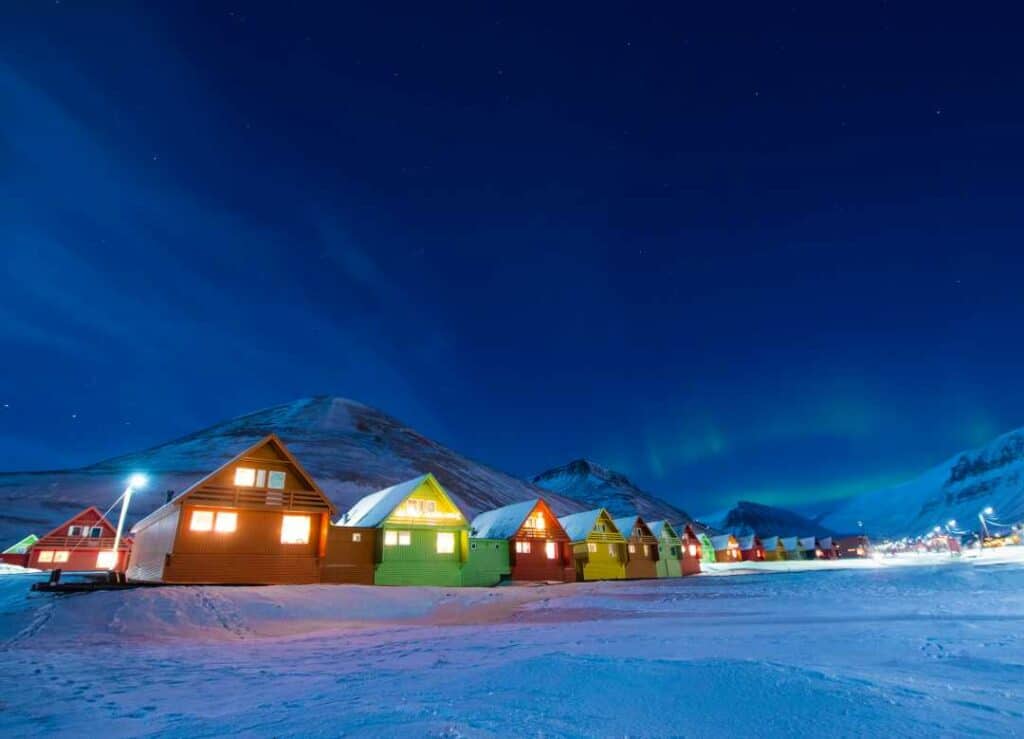 Longyear byen, the capital in Svalbard, in the winter. The snowy ground, the mountains in the back and the dark sky all have shades of blue - the winter colors of the Arctic. In the foregorund are the typical colorful wooden houses of Svalbard, red, green and yellow, lit up from the inside. There is a whiff of northern lights in the distance. 