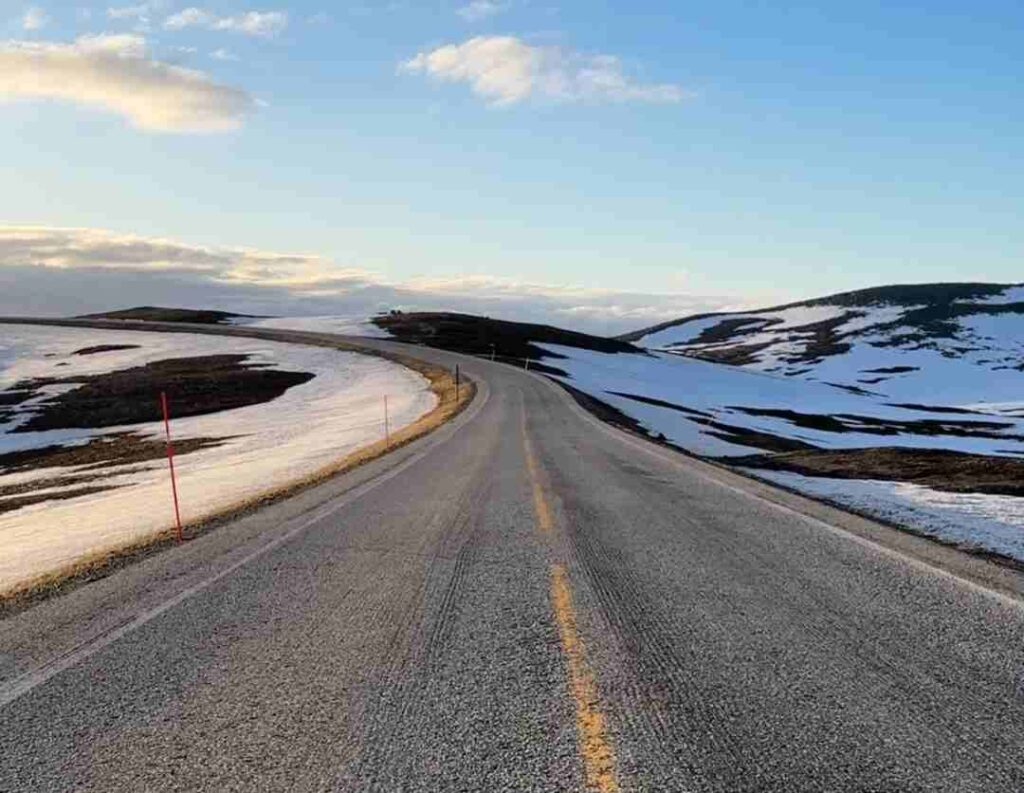 A wide road stretching into the mountain plains with patches of snow on the sides under a blue sky