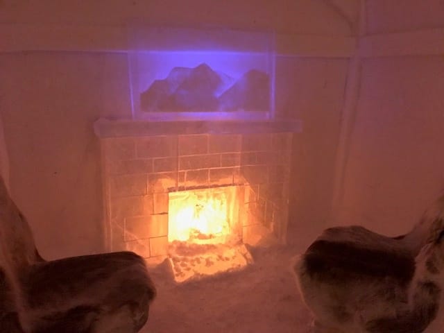 A fireplace inside the ice hotel - made entirely of ice, with ice chairs covered with raindeer furs in front of the fire, the room glowing in a misty atmosphere. 