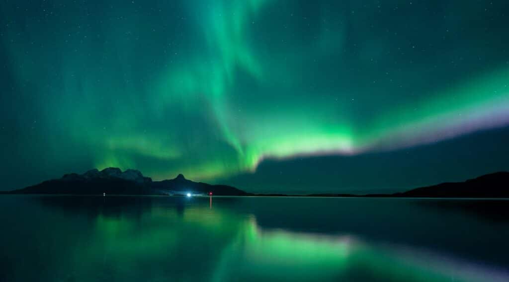 The Aurora Northern Lights outside Bodo, Norway, the greenish, purplish, with some blue in waves of light across the dark night sky, reflected in the bland dark water in the foreground. 