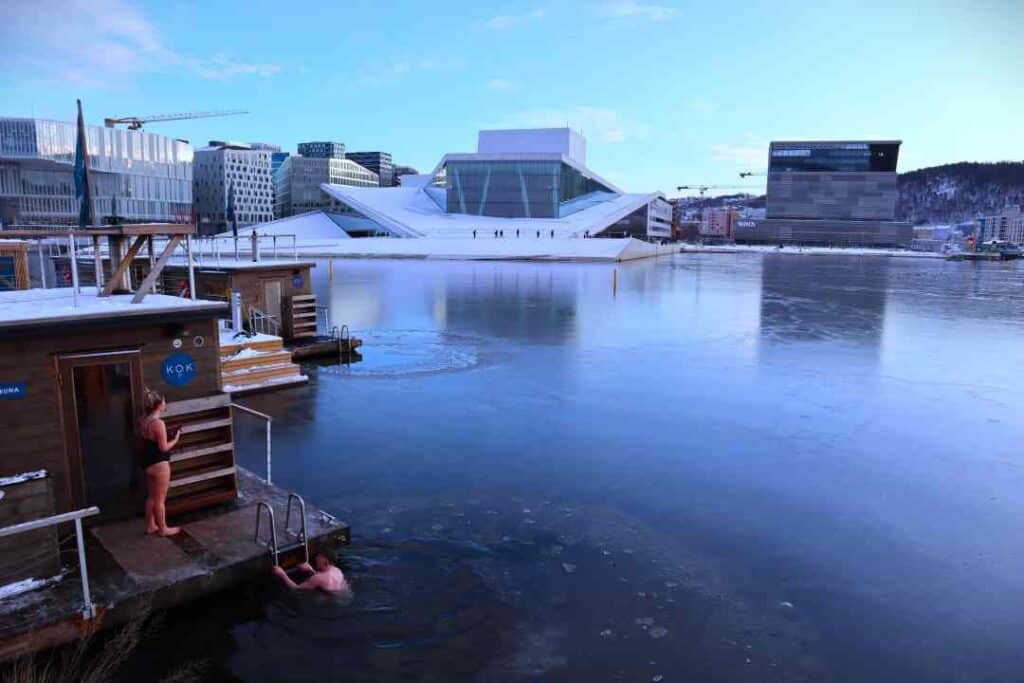 A white marble building in modern architecture with slopey roof where people are walking, across a ice layered fjord, with saunas on the near side, under a pale blue winter sky. The Oslo opera house. 