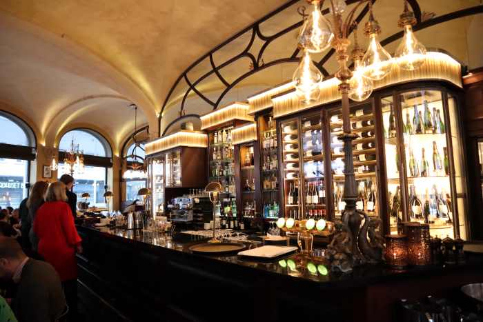 Is Oslo worth visiting? The Theater Cafe in Central Oslo, with lots of guests around the bar area, with warm golden lighting and shiny shelfs behind the bar under elegant decorated ceilings. 