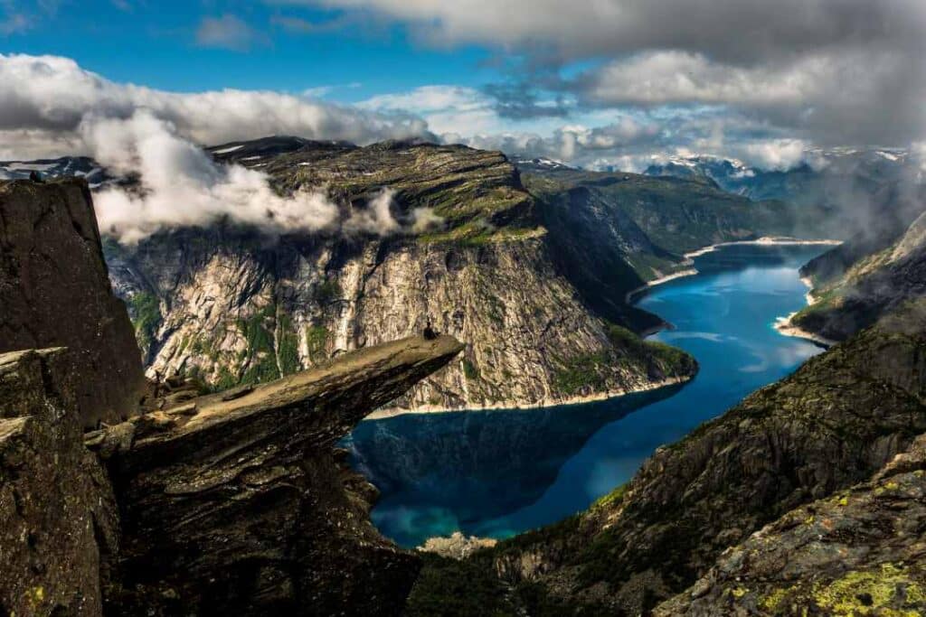 A dramatic cliff high on a mountain top over a narrow, deep blue fjord surrounded by steep mountains at Trolltunga, Norway.