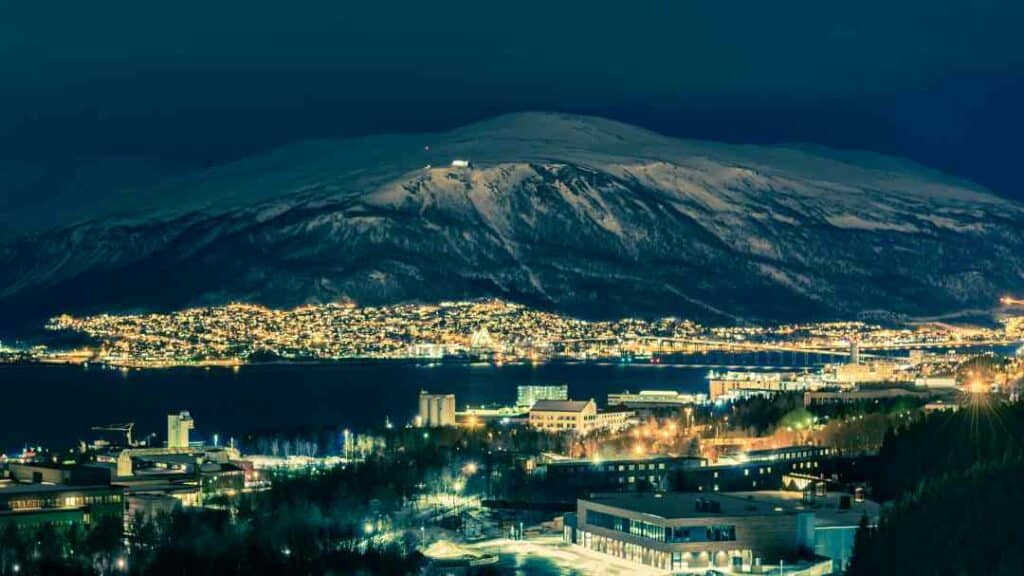 Tromsø on a dark winter night, with glowing lights from the city under the dark sky and snowy mountain across the fjord. You see the Tromsø bridge connecting hte mainland, and the Ishavskatedralen Cathedral looking like a pyramid on the other side of the water. 