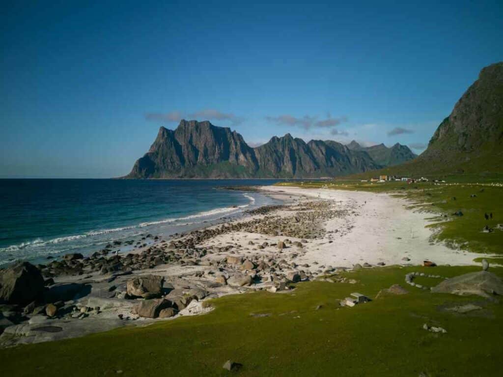 Uttakleiv Beach in Lofoten on a clear sommer day, with the white sand below the green grassy fields with a few scattered farms in the distance. The backdrop is the vast blue sea, and the steep mountains in the distance. 