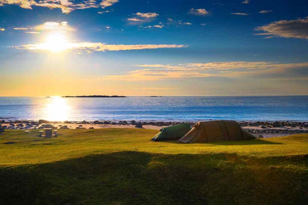 Uttakleiv beach in Lofoten on a summer day with a deep blue sunny sky, green grassy fields by the golden sandy beach, and two tents set up in the middle of this stunning nature