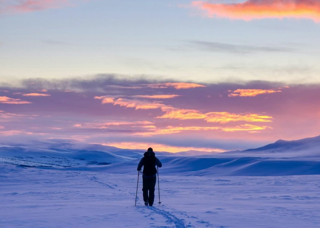 A person skiing alone into a winter wonderland surrounded by white snow covered plains under a pale blue sky glowing from the sun in the horizon