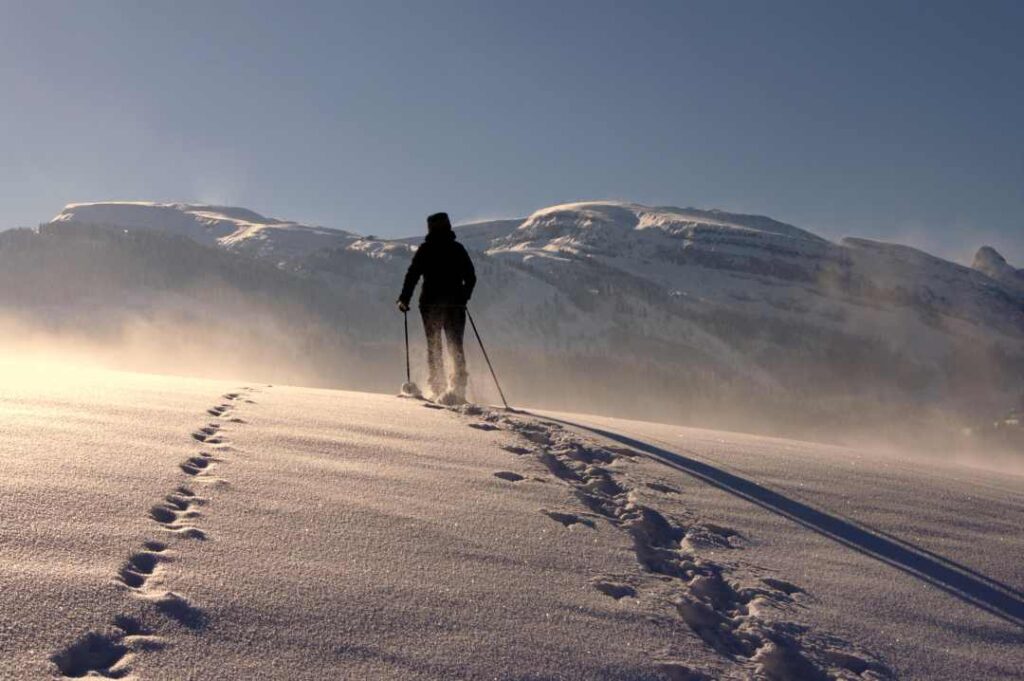 Solo skier cross country across a snowy mountain plain, white snow flying in the wind over the plains, blue skies above, and the light is glowing from the winter sun outside the picture.