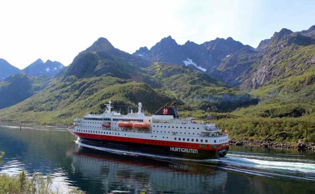 The Hurtigruten coast line ship in a norwegian fjord in summer, surrounded by green mountains with patchy snow 