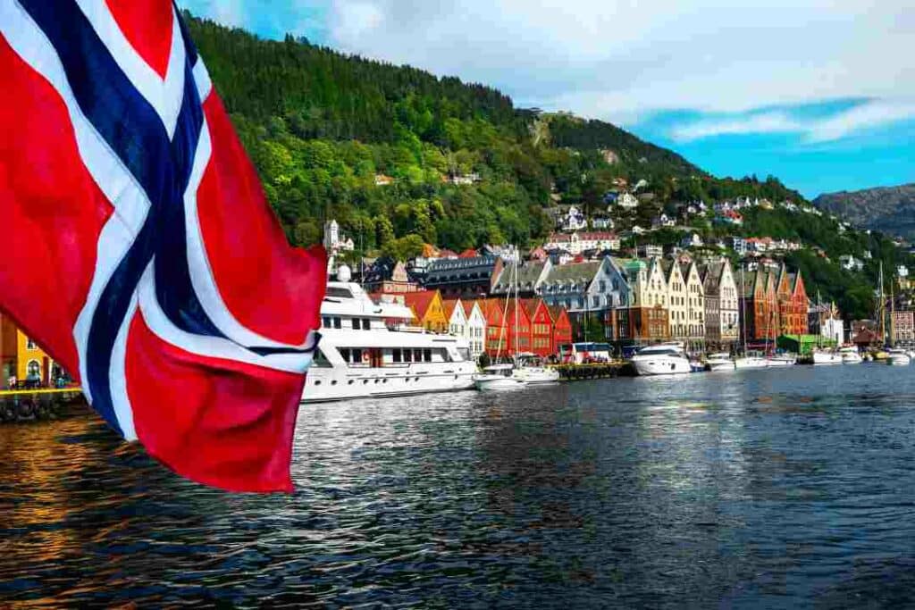 The dark blue waters of Bergen Harbor in the summer seen from a boat, with a norwegian flag in the aft. You see the docks with numerous boats, the famous particular houses and architecture of the Bergen Wharf, and the green hills with houses climbing along in the background. 