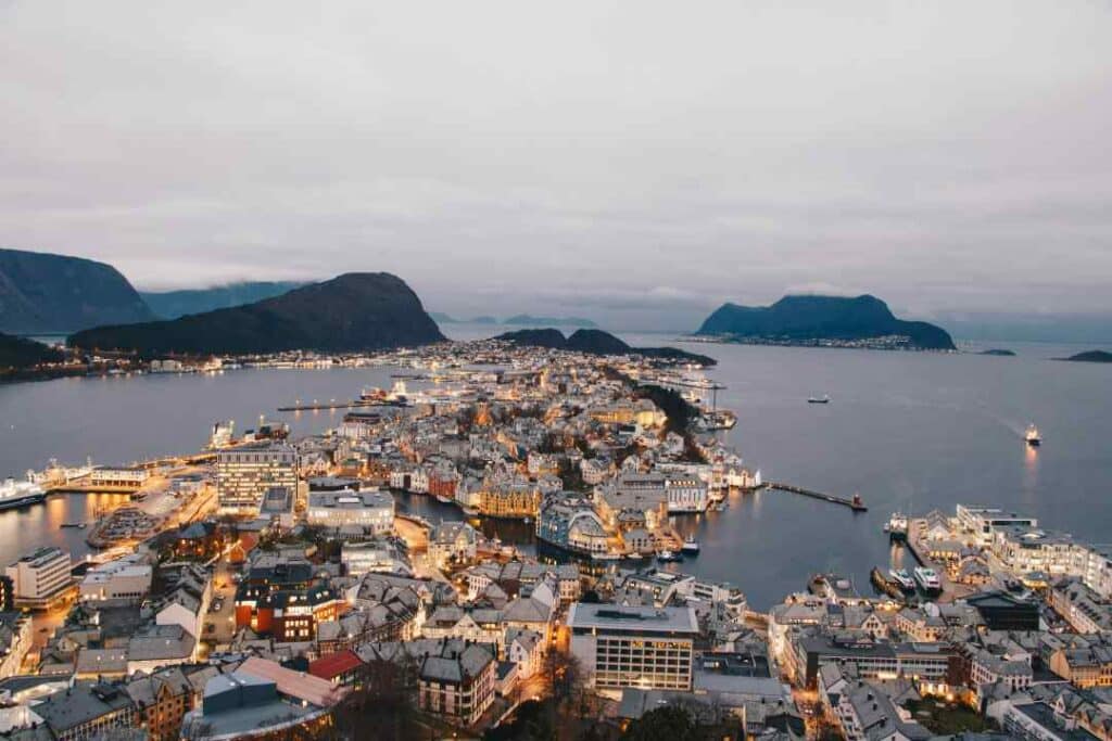 The town of Ålesun at dusk, with lights coming on in the city that is situated right by the fjord shores with stunning views of the water and the mountains in the distance