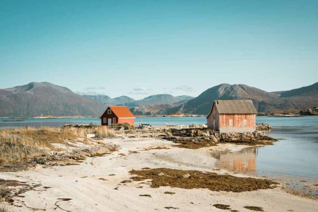 A white sandy beach outside Tromsø, Norway on a sunny summer day, with two red old wooden cottages next to the blank water