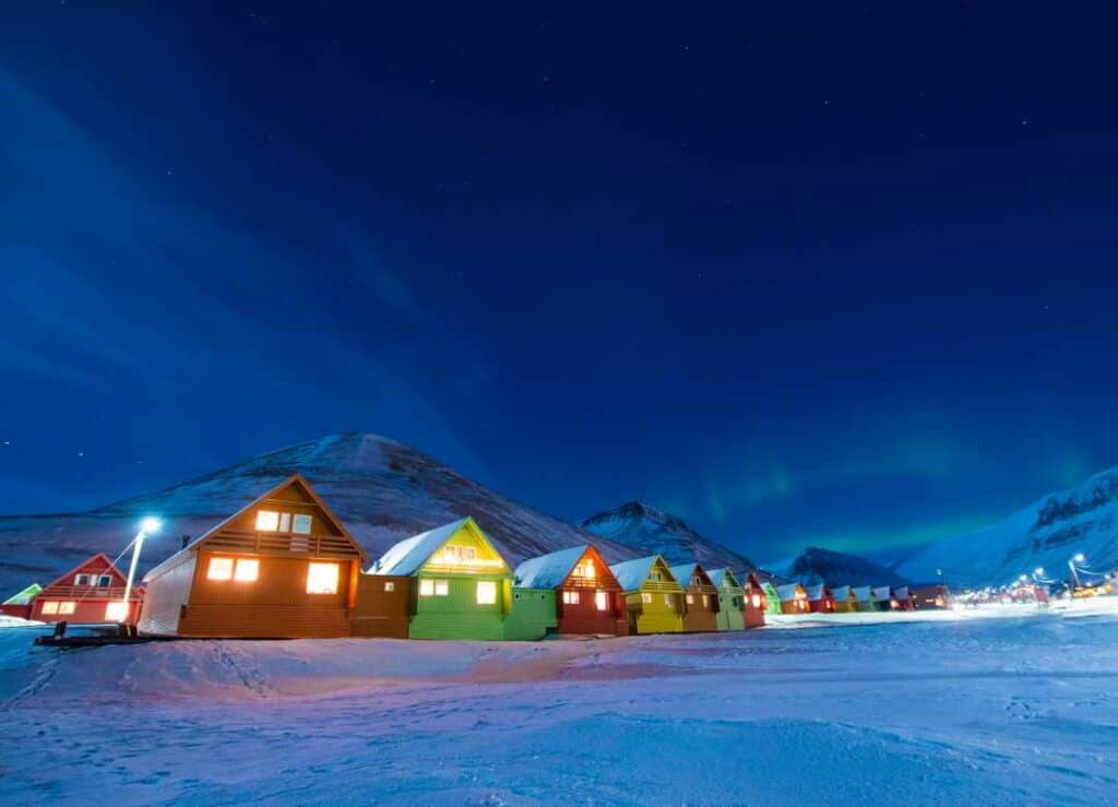 The dark blue polar night in Svalbard Norway, the sky is deep blue, the snow is white with a blue shimmer, the mountains white with a blue shimmer, and the colorful houses lit by warm lights