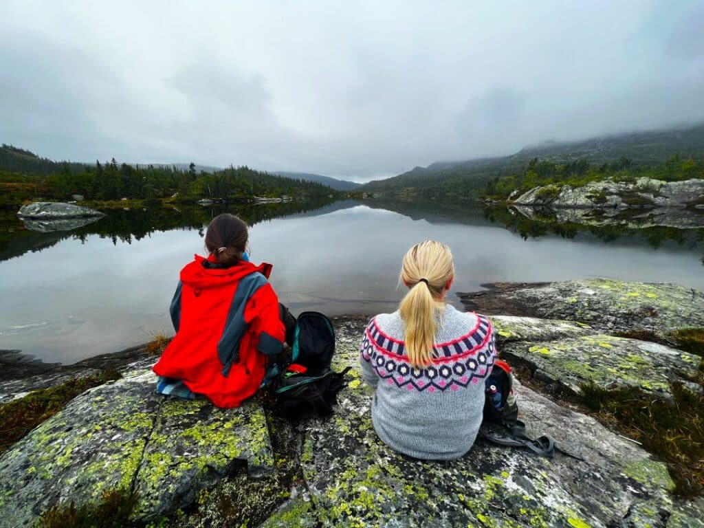 Two girls sitting by a silvery mountain lake in Norway in the winter, surrounded by misty air and green hills