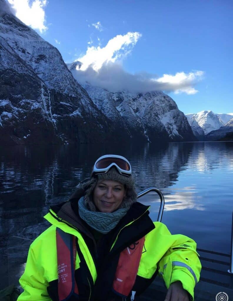 A lady dressed in a survival suit with goggles and hat on a RIB with the blank dark blue fjord behind her, and steep snow covered mountain sides hovering over the fjord under a blue sky