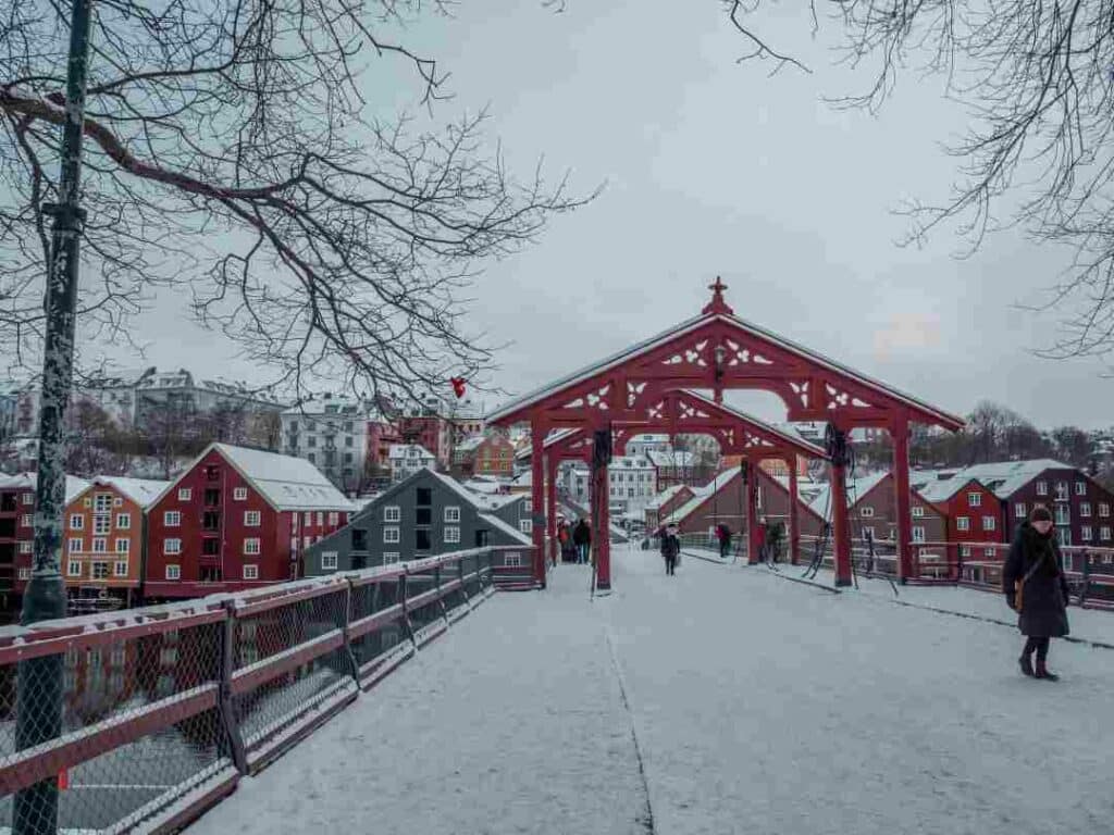 An old decorated bridge in the winter, covered in snow under trees with no leaves, and a red wooden archway covering the bridge in the middle