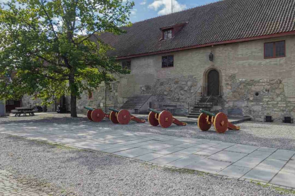 Old cannons with wooden brown wheels in an old stone courtyard surrounded by stone houses on a summer day with green leaves on the trees