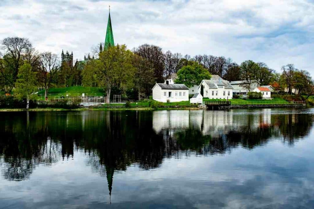 A dark blue wide river reflecting the white wooden houses on one side, sitting in the middle of a green park with trees and grassy fields. A church spear is seen above the treetops under the cloud dotted sky. 