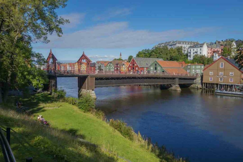 An old bridge over a dark blue river with green trees and grass on one side, and old city buildings on the other, under a blue summer sky