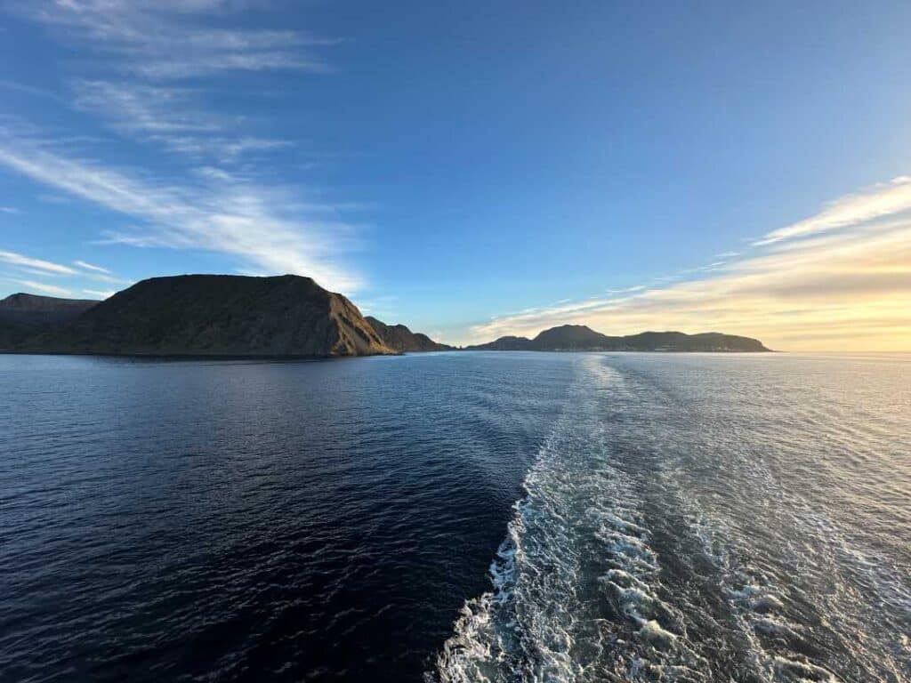 The aft whitewater trailing a ship sailing on dark water with a low glowing sun in the distance over majestic mountains