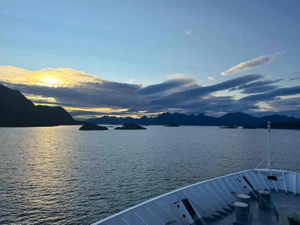 Norwegian coastal voyage: Small islands in the distance in front of a pale sunset on a blue sky, the photo is taken from the white bau of a ship sailing in polar waters