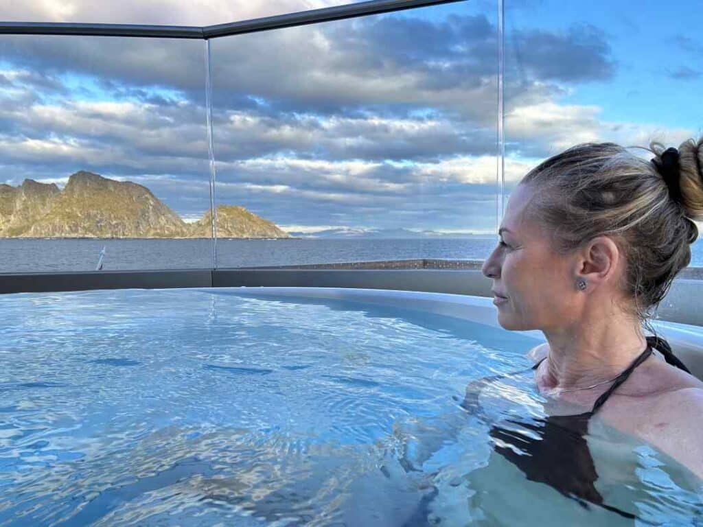 Lady sitting in a hot tub on the deck of a ship, gazing at the barren mountains in the distance under blue skies with dotted clouds