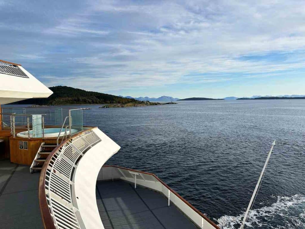 The bau area of a ship at sea, with a hot tub on the side, and mountainsin the distance beyond a fjord with dark blue waters under the pale blue sky