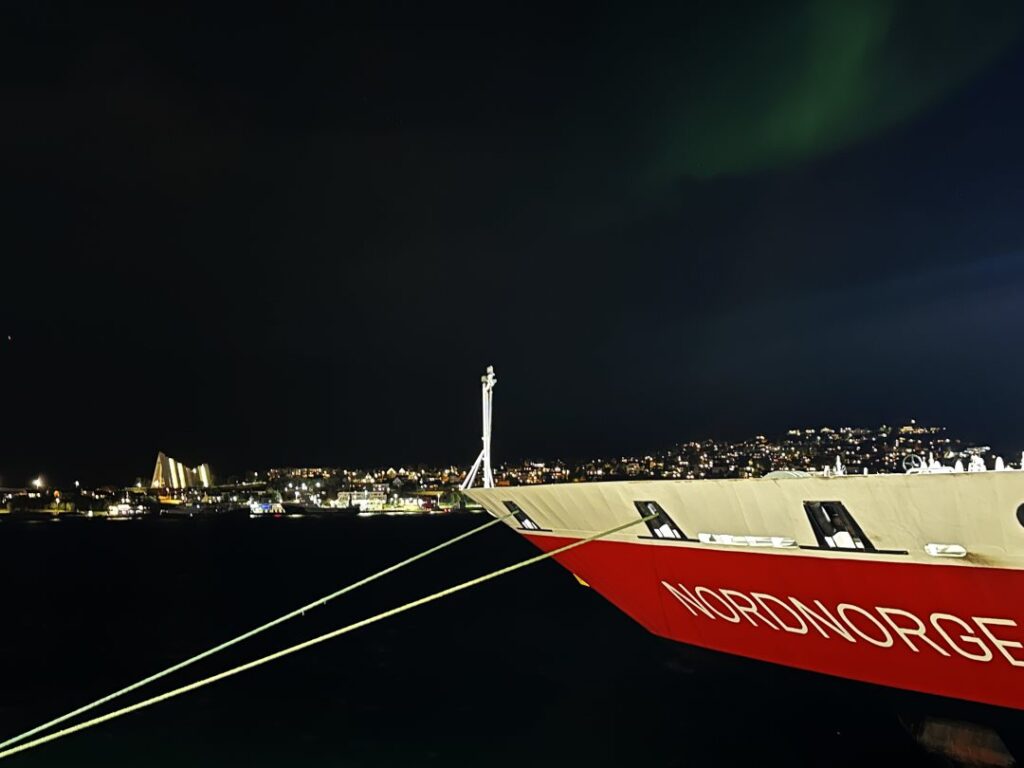 A white and red ship bau docked at night on dark water under a dark sky with a whiff of northern lights in the dark sky above and a glowing town in the background