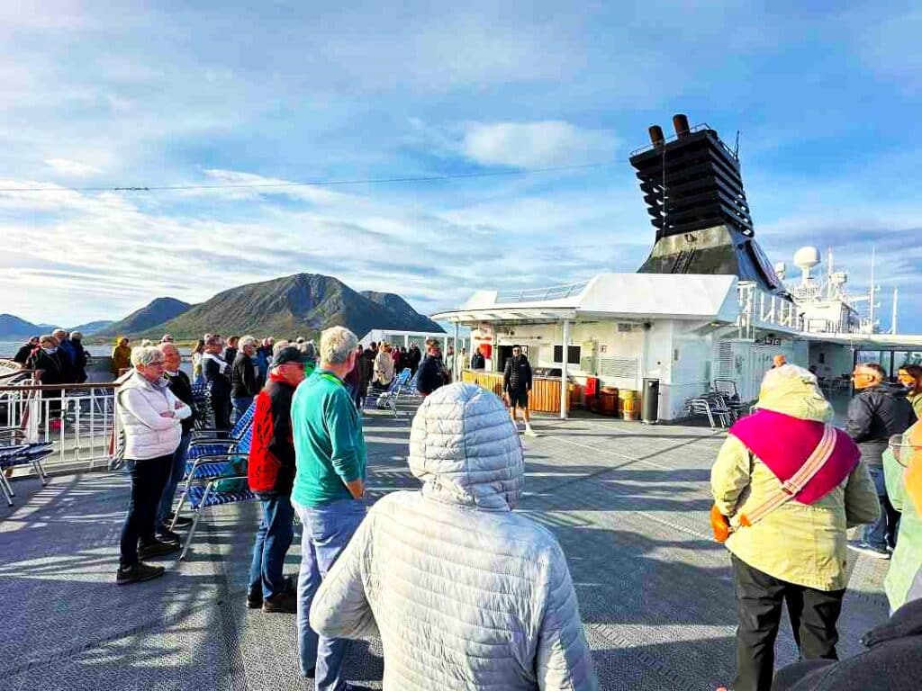 a group of people on the deck of a ship wearing winter clothes, listening to a guide telling stories about the Arctic region surrounding the ship 