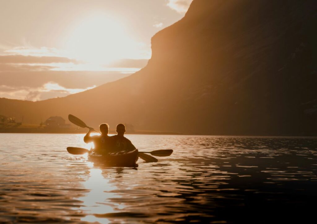 Two people kayaking in a fjord below mountains under the midnight sun